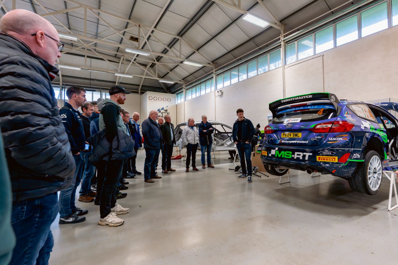 Rally sponsors listening to a Jon Armstrong explaining a rally car in the high-ceiling M Sport workshop.