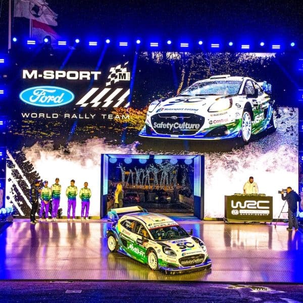 Jon Armstrong and M-Sport Ford World Rally Team launch stage at Rallye Monte Carlo, Monaco Harbour, with a Ford Puma Rally1 on the platform under bright lights and a giant screen showing the car and team branding.