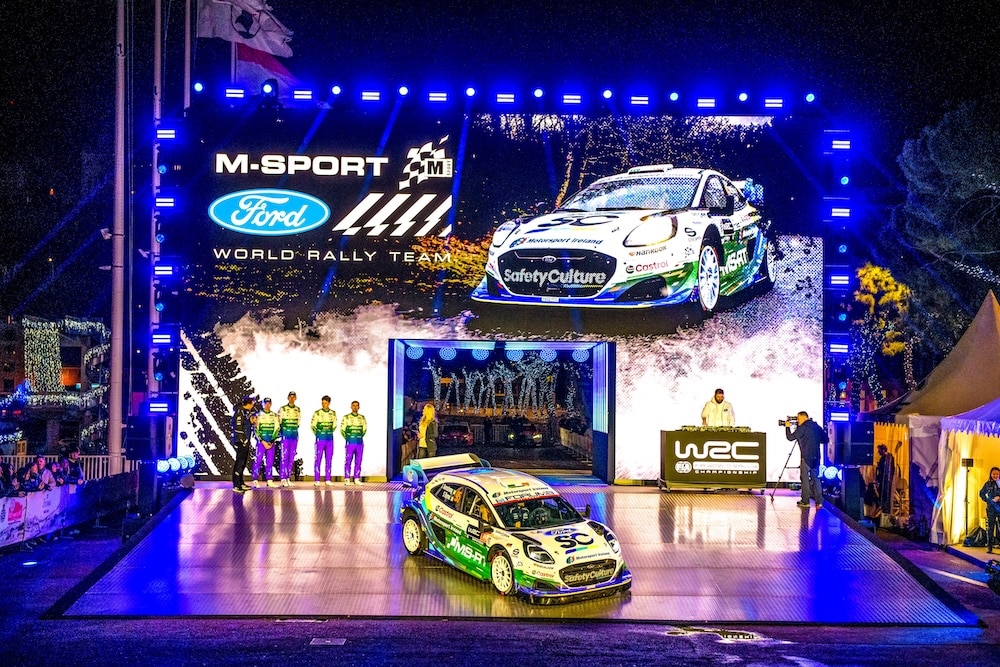 Jon Armstrong and M-Sport Ford World Rally Team launch stage at Rallye Monte Carlo, Monaco Harbour, with a Ford Puma Rally1 on the platform under bright lights and a giant screen showing the car and team branding.